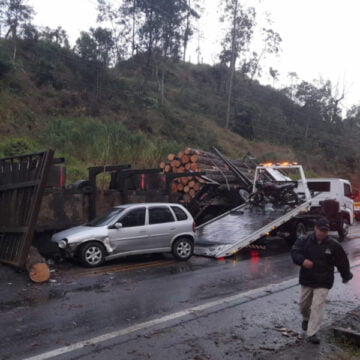 Caminhão com carga de madeira tomba e carro bate em toras caídas na pista